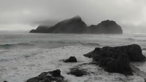 Aerial-forward-over-sea-with-big-rock-shrouded-in-mist-on-background,-Ponta-da-Calheta