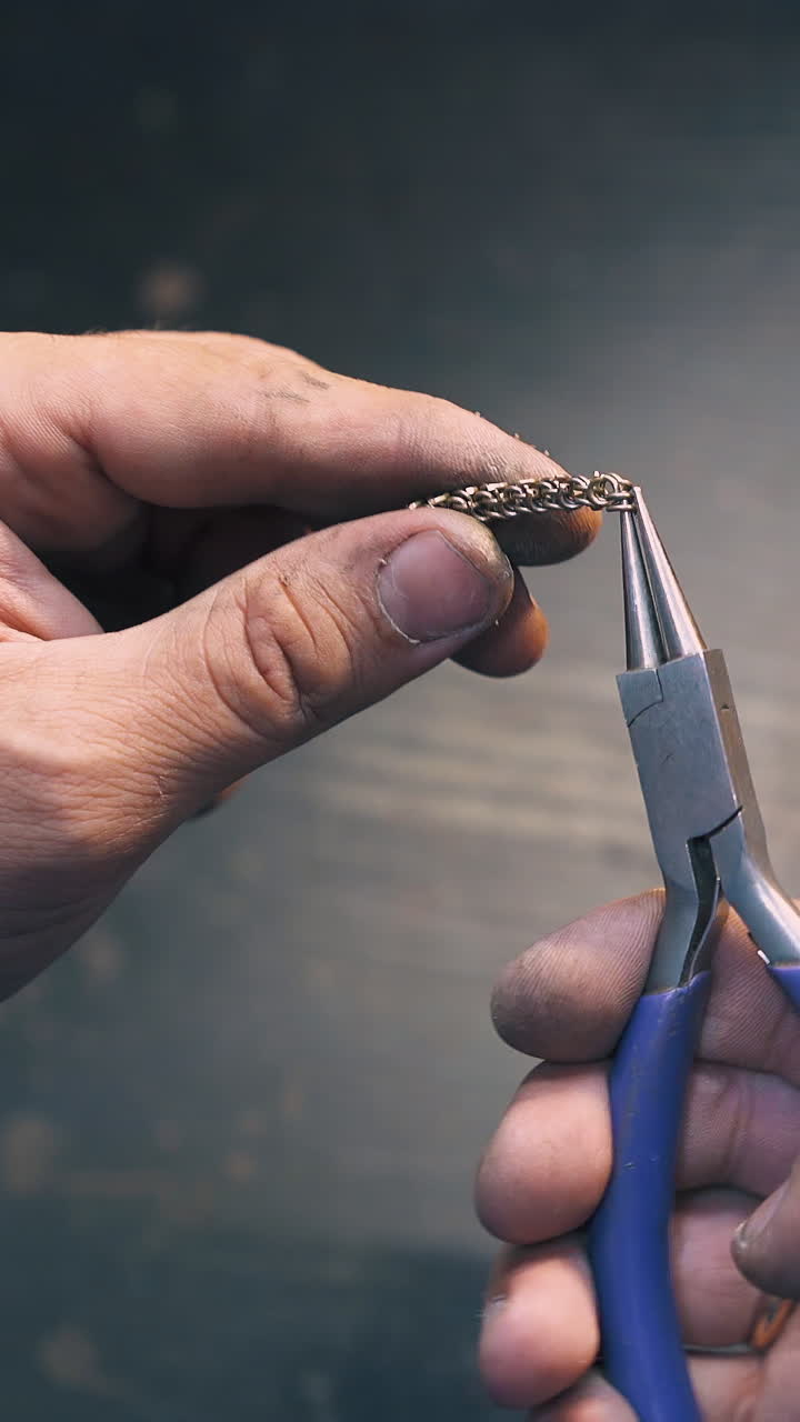 CU shot: young goldsmith connects little links to chain above dark wooden table in light jewelry craft workshop close view