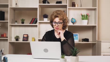 Una-Mujer-Mayor-Con-Gafas-Está-Chateando-Por-Video-En-Una-Computadora-Portátil-Mientras-Está-Sentada-En-La-Mesa