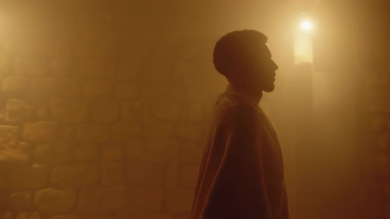 Una coppia ammantata si muove attraverso una camera di pietra chiusa, con una silhouette che guida una seconda figura accanto a pareti ruvide, mentre una calda retroilluminazione, foschia e ombre morbide creano un'atmosfera cinematografica misteriosa.