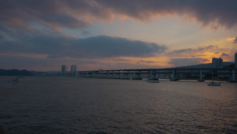 silhouetted travel tour yachts cruising along gwangan bridge in busan, dramatic sky during golden sunset in busan