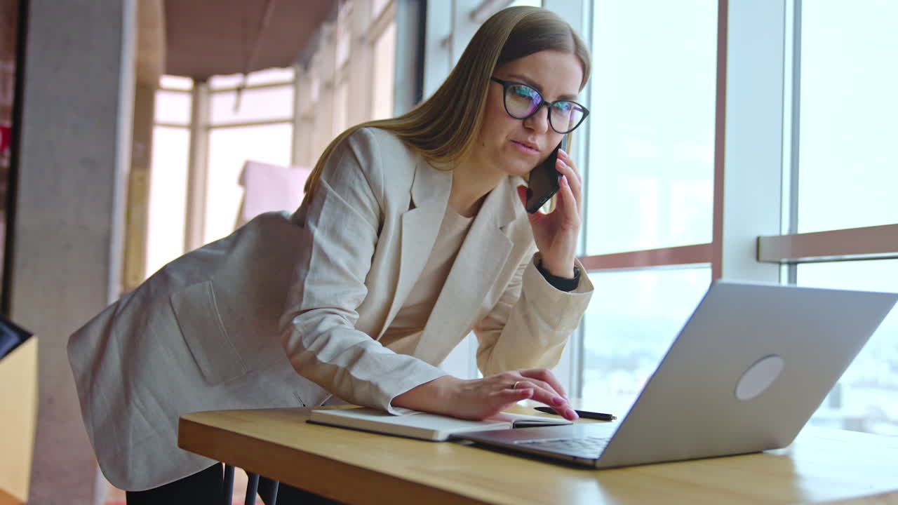 Lady in light suit standing leaned over the desk. Smiling woman searches something on laptop and speaks on the phone.