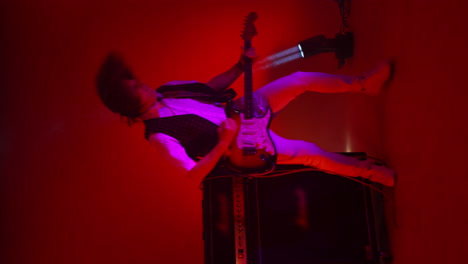 Vertical-video-crazy-male-rock-musician-playing-guitar-in-the-Studio-in-the-light-of-spotlights-and-smoke-dancing-and-jumping-in-the-Studio-shaking-her-hair-and-head