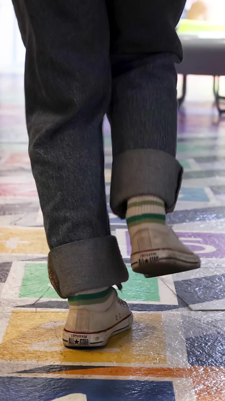 Un adulto che indossa scarpe casual e jeans salta lungo una colorata griglia per la campana in un parco giochi interno ben illuminato, ripreso da una prospettiva posteriore bassa.
