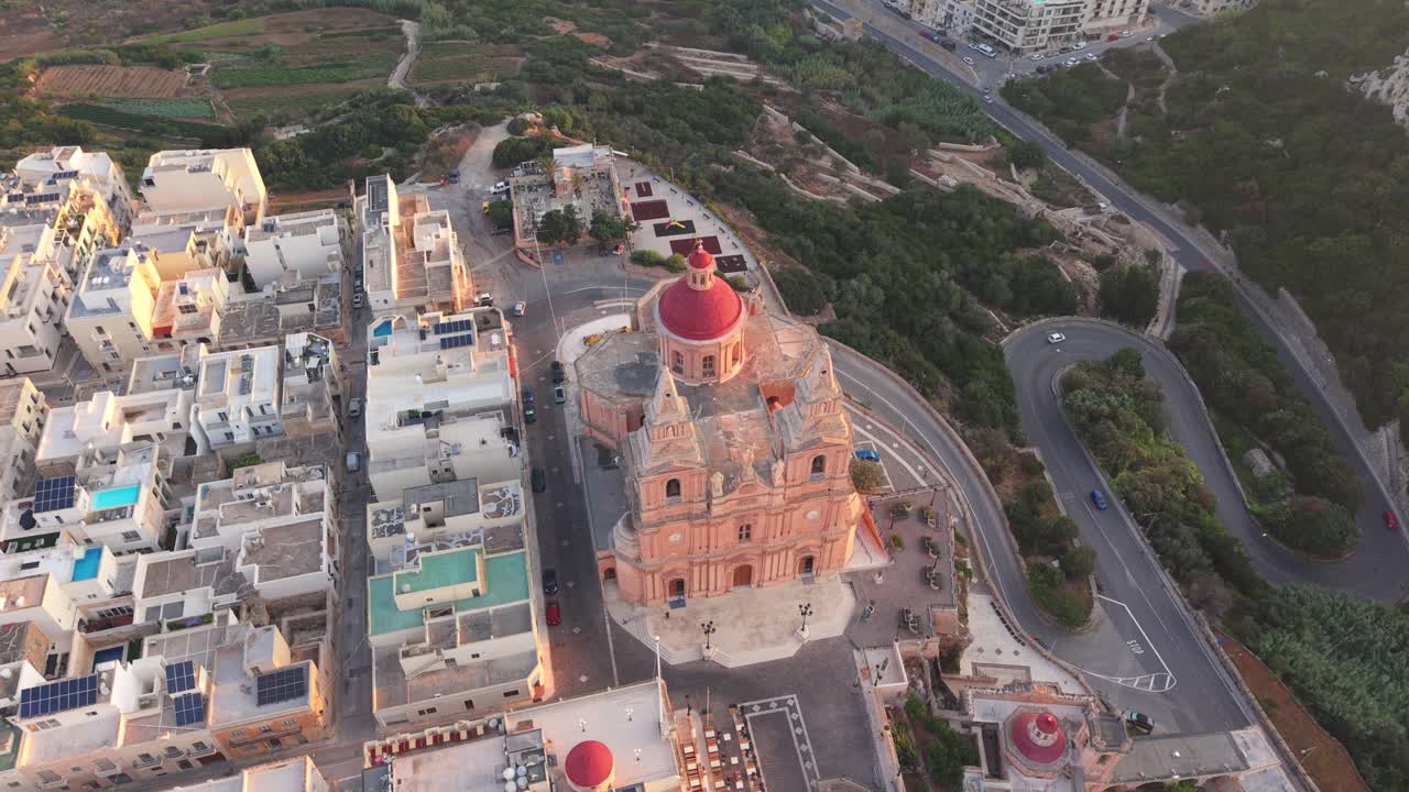 An aerial view of the Mellieha Parish Church in Malta. The baroque-style church with its red dome stands out among the surrounding town buildings with the countryside in the background