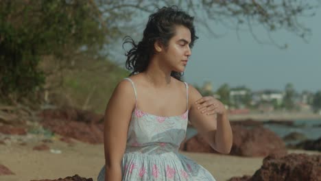 An-Asian-girl-in-a-floral-dress-stands-on-a-rocky-beach,-shading-her-eyes-with-her-hand-as-she-gazes-into-the-distance