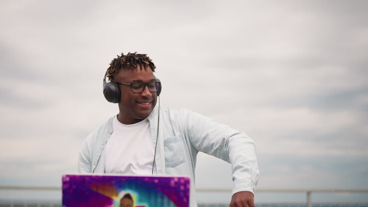 DJ sorridente con cuffie e controller che mixa beat elettronici soulful su una console portatile vicino a un lungomare nuvoloso, primi piani di ritratti catturano un'atmosfera rilassata, ritmo espressivo e performance intima.