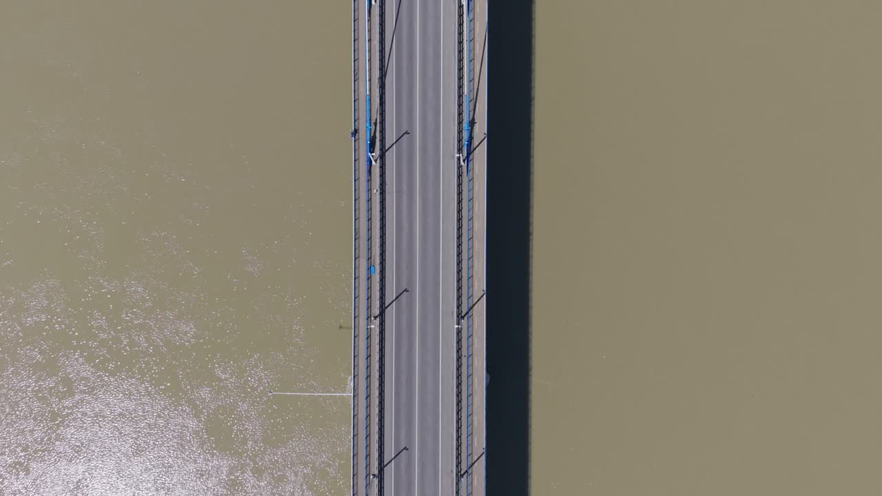 Aerial view of vehicles traveling across a bridge over a wide brownish river. The symmetrical composition and the stark contrast between the vehicle and the water create a striking visual from above