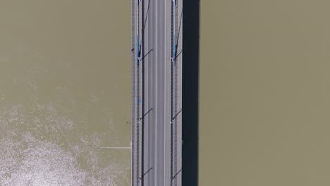 aerial view of vehicles traveling across a bridge over a wide brownish river. the symmetrical composition and the stark contrast between the vehicle and the water create a striking visual from above