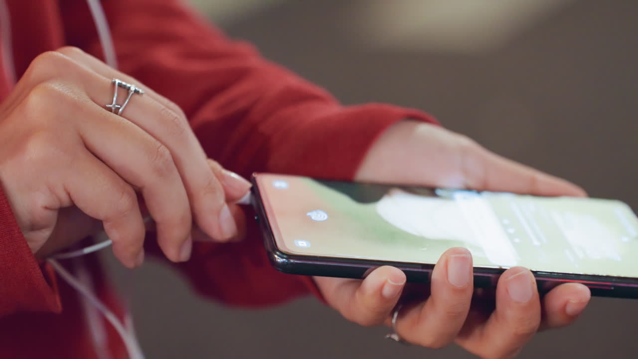 Mani in primo piano che tengono uno smartphone e auricolari, con una felpa rossa, mentre toccano il touchscreen per navigare nella playlist; cavo di ricarica e anelli d'argento visibili, luci sfocate della città con effetto bokeh, atmosfera da pendolarismo serale, manicure casuale.