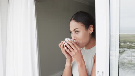 Mujer-Birracial-Pensativa-Mirando-Por-La-Ventana-Bebiendo-Té-En-Casa,-Cámara-Lenta