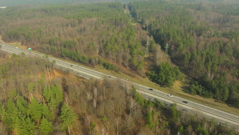 aerial shot capturing a highway slicing through a lush forest in gdynia dąbrowa, with vehicles in transit and power lines paralleling the road, illustrating infrastructure within natural surroundings