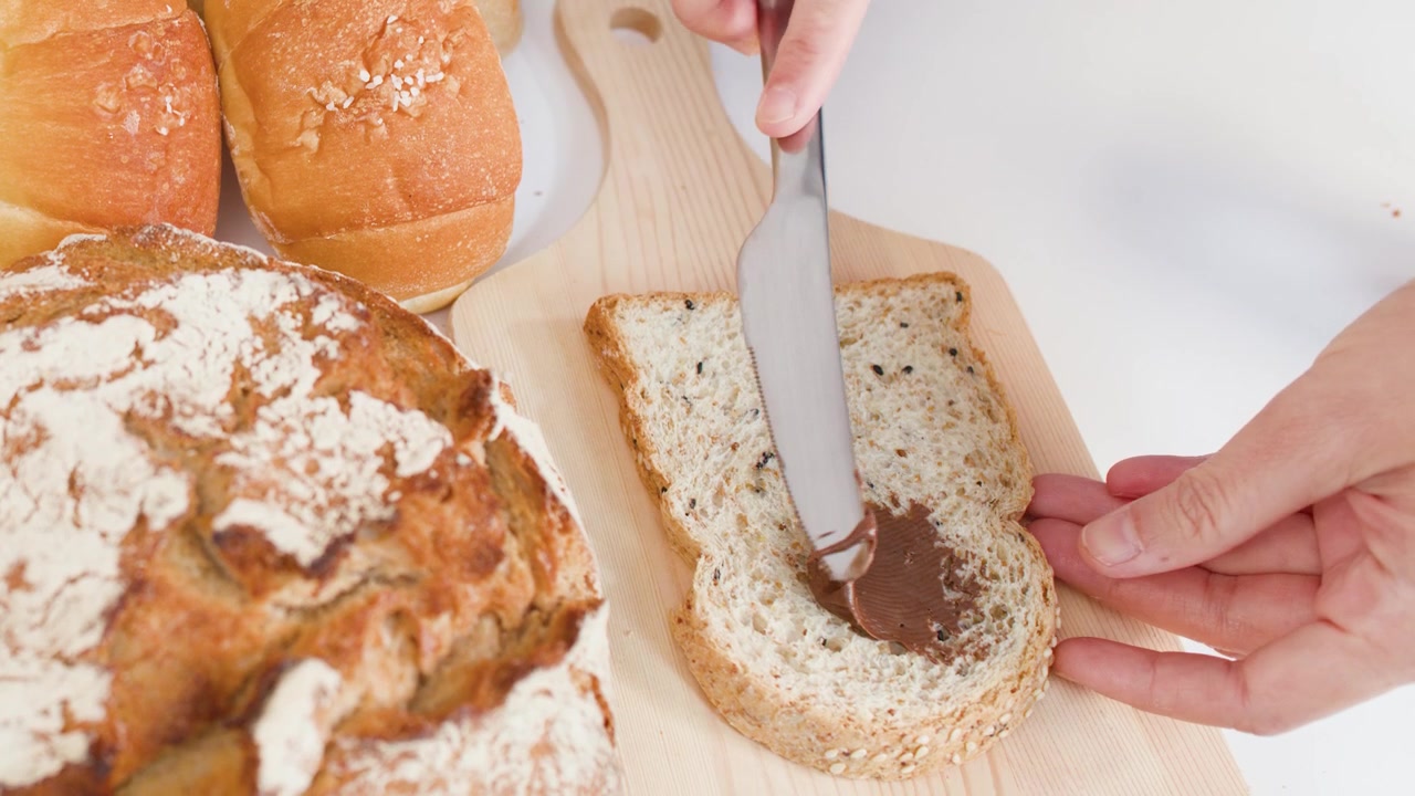 Primo piano di mani che spalmano crema spalmabile al cioccolato e nocciole su pane a lievitazione naturale su un tagliere di legno.