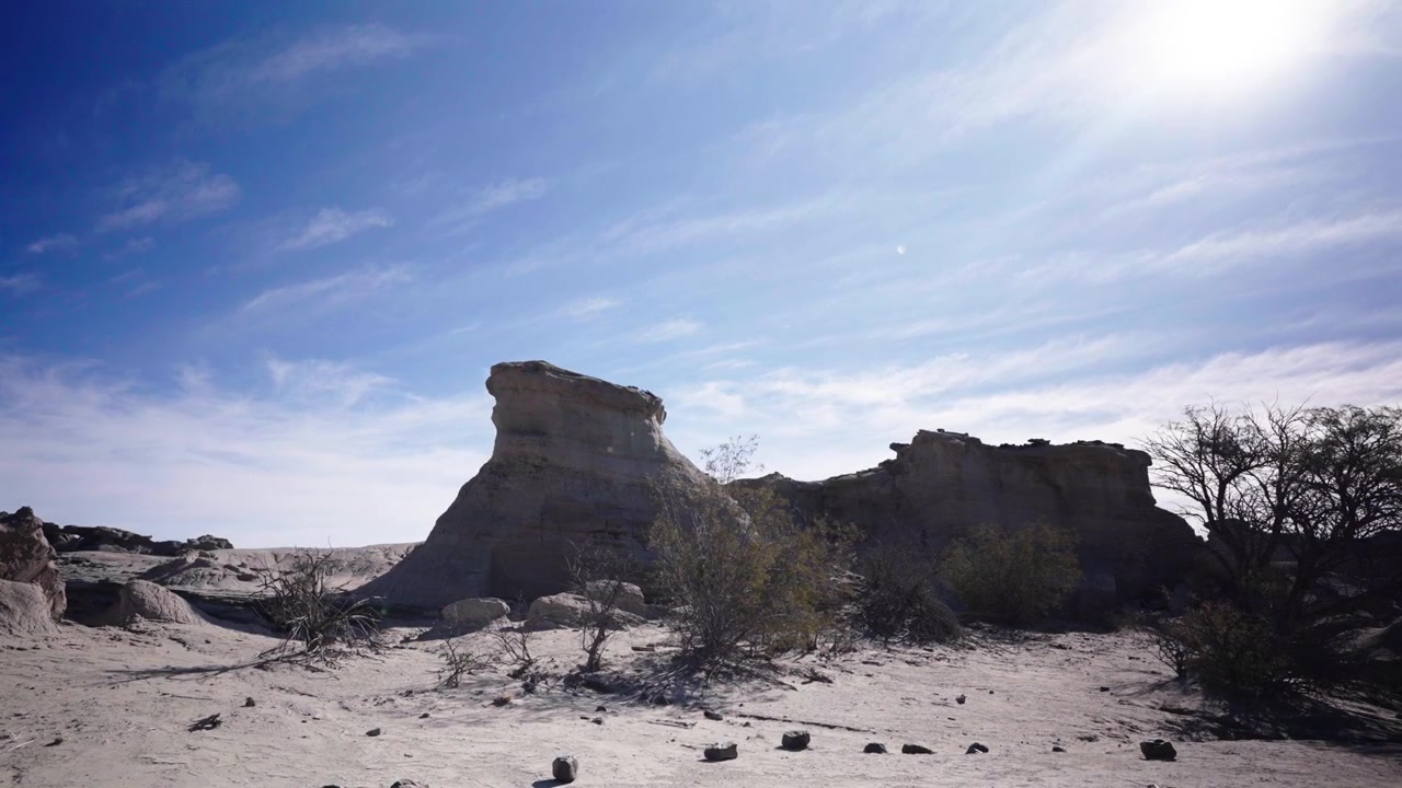 Formazioni rocciose di arenaria illuminate dal sole si ergono tra rari cespugli desertici nel Parco Provinciale di Ischigualasto, Argentina, sotto un cielo azzurro brillante con nuvole sparse
