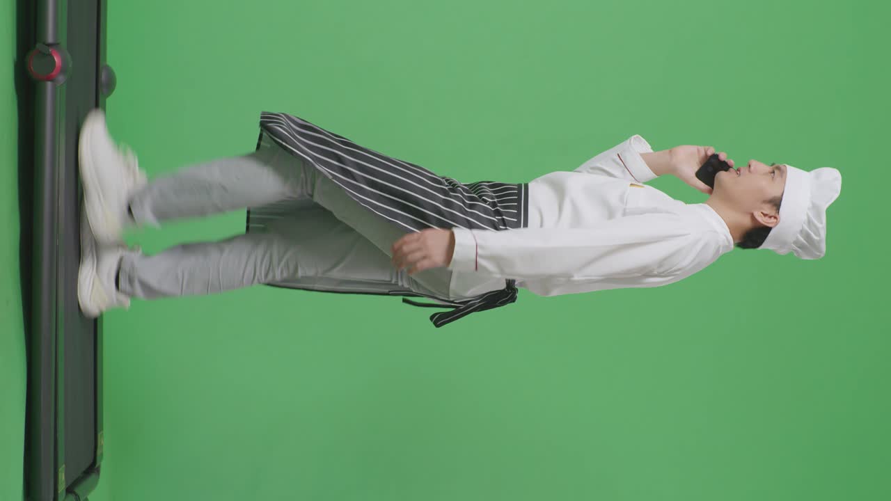 으로 보이는 아시아 남성 셰프가 녹색 스크린 배경 스튜디오에서 는 동안 스마트폰으로 이야기하고 있습니다.