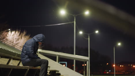 a man wearing a hooded jacket sits alone on bleachers at night, illuminated by streetlights, with a dimly lit building in the background