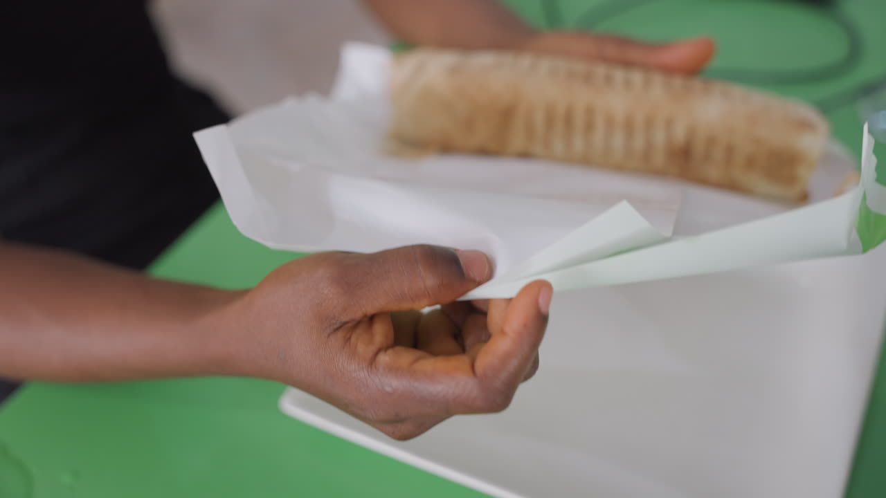 Black hands wrapping sandwich on plate, home cook packing grilled wrap into paper for romantic picnic date, green table surface, white square plate, napkin folding, intimate gentle gestures, cozy