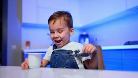 funny toddler boy holding a yogurt cup in mouth and spoon in hand. some dairy spots are on kid's face.