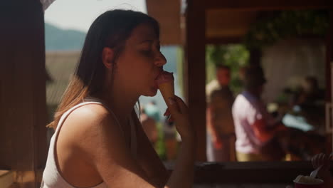 languid woman bites tasty ice cream in wafer cone sitting at table in cafe slow motion. lady enjoys frozen dessert resting on restaurant terrace