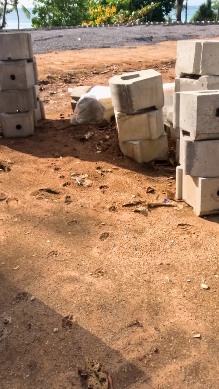 Cinder blocks stacked on sandy ground near a Phuket beach, captured in bright daylight with shadows and ocean in the background