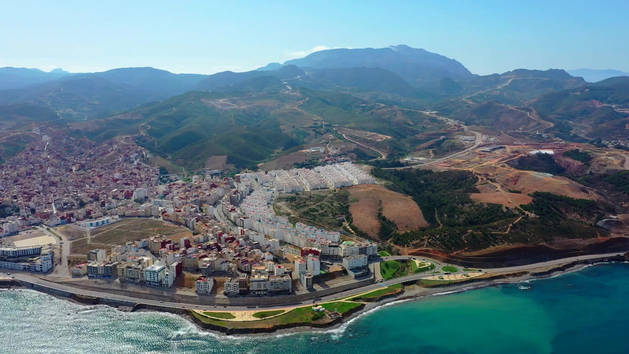 Vista aerea panoramica di una città costiera che si estende tra le acque turchesi del Mediterraneo e i Monti del Rif, rivelando lo spettacolare paesaggio che va dal mare alla montagna del Marocco settentrionale