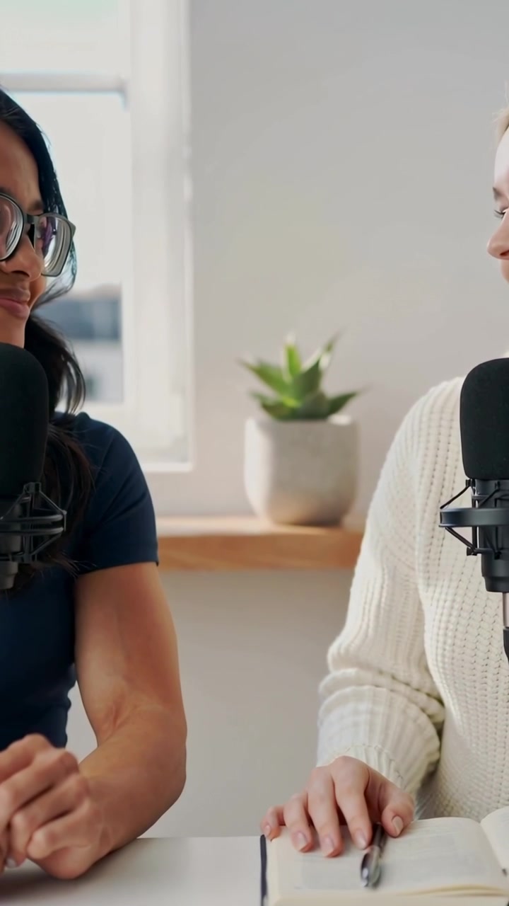 Video verticale: Due conduttori uno di fronte all'altro parlano nei microfoni durante una registrazione in studio, con maglia blu navy e appunti.