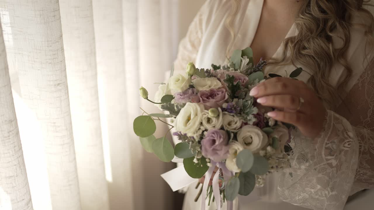 Sposa in vestaglia di pizzo tiene un delicato bouquet floreale vicino alla finestra