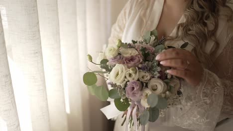 bride in lace robe holds a delicate floral bouquet near the window