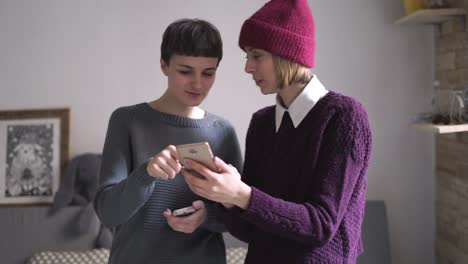 Two-young-woman-looking-mobile-phone-standing-in-room.-Woman-holding-smartphone