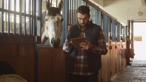 man is using a tablet computer next to a horse in a stable.