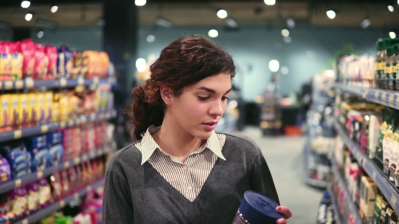 una joven morena toma una botella de café del estante de la tienda de comestibles, la abre para olerla y pone la botella en el carrito de compras.
