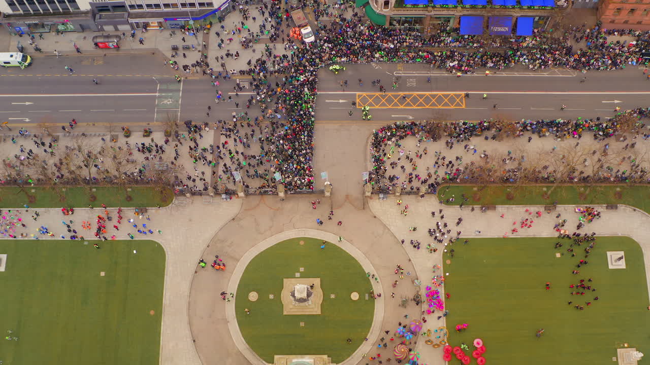Top-down aerial pan over St. Patrick’s Day performers on Town Hall grass with awaiting crowd