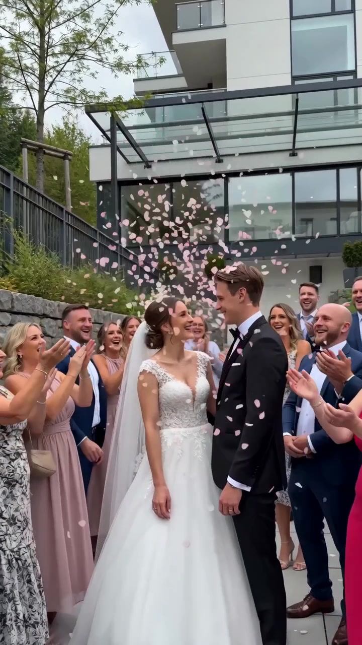 Sposi stanno insieme mentre gli invitati acclamano con le mani alzate, mentre petali rosa cadono intorno a loro davanti a un moderno edificio con facciata in vetro.