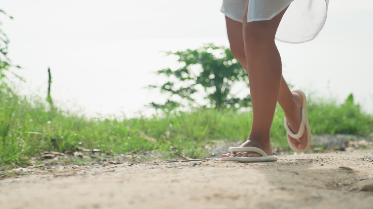 Soft sunset legs walking along island shore, golden light and gentle breeze, sandals tapping pebbles and gravel, slow reflective steps, intimate evening mood of seaside escape and quiet romance