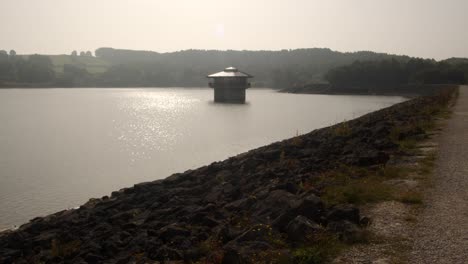 Toma-Extra-Amplia-De-La-Torre-De-Válvulas-De-Agua-De-Carsington-Water,-La-Torre-De-Extracción-Y-Las-Rocas-De-La-Presa-Con-El-Sol-Detrás