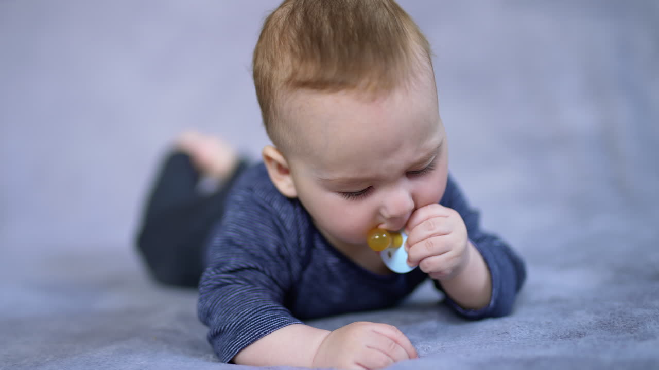 Cute adorable baby boy dealing with a pacifier. Beautiful infant child chewing the soother and looking aside. Close up. Grey blurred backdrop.