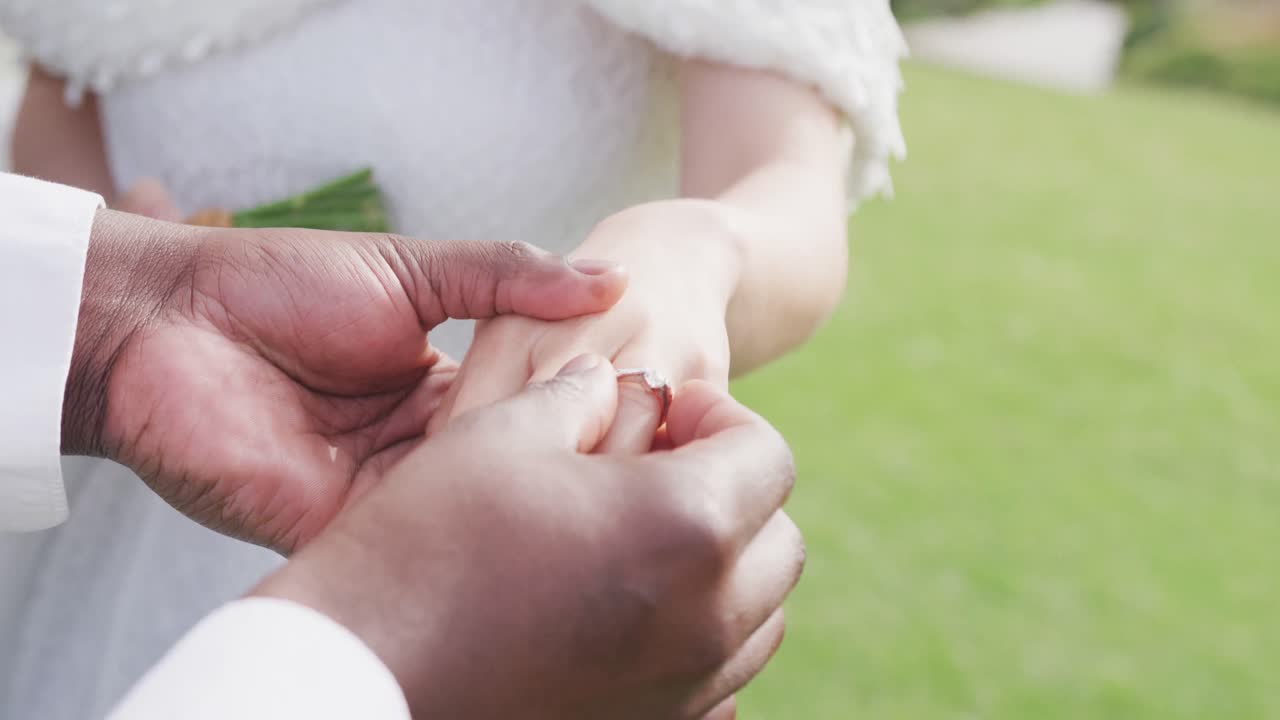 Video of hands of diverse bride and groom, groom putting ring on bride's finger at outdoor wedding