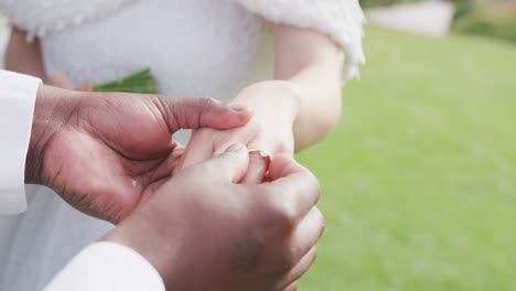 Video-of-hands-of-diverse-bride-and-groom,-groom-putting-ring-on-bride's-finger-at-outdoor-wedding