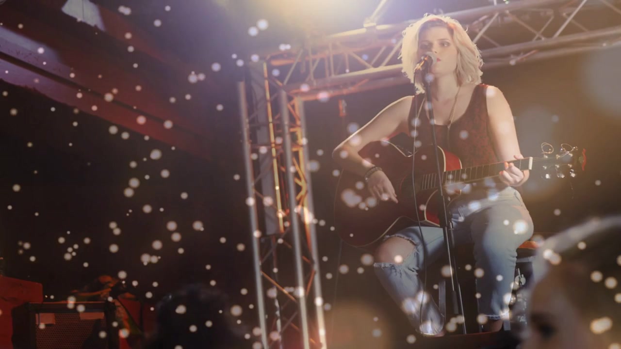 Kamera, oturan kadın müzisyene doğru kayıyor; gitar çalıyor ve bokeh efektiyle müzikseverlerle etkileşim kuruyor.
