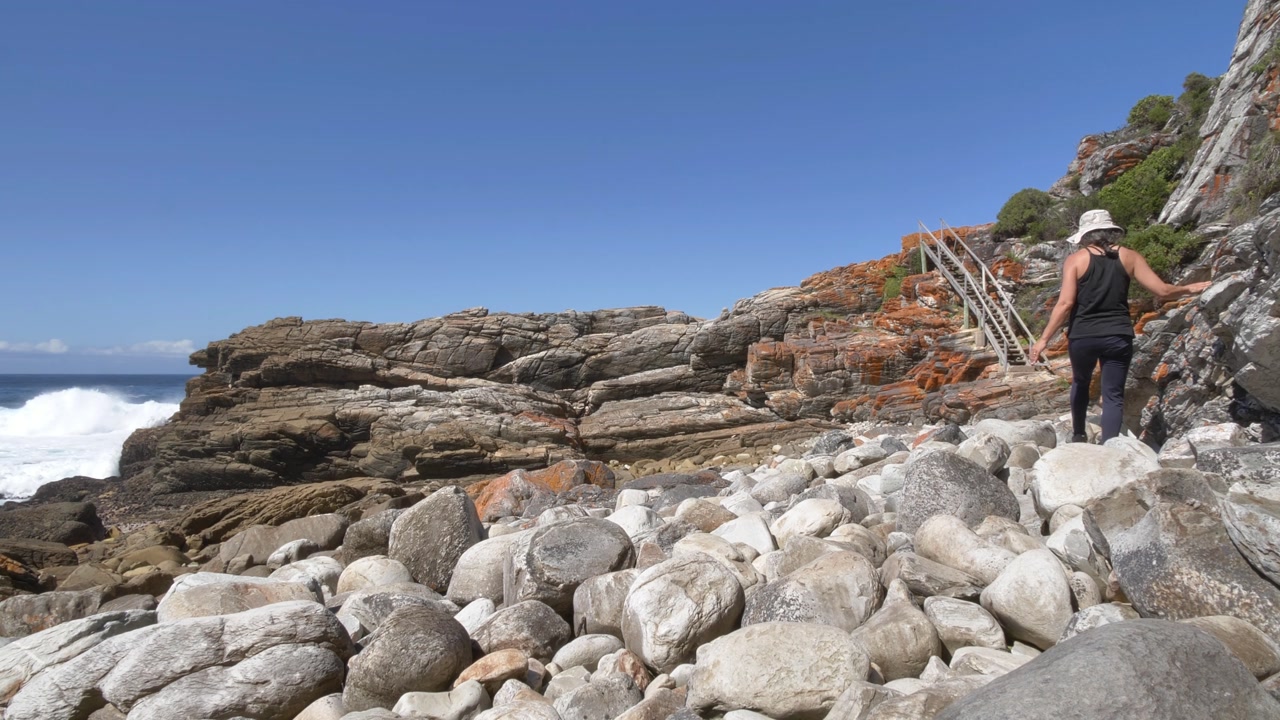 Two women navigate through rocky terrain towards a wooden staircase along a rugged coastline, with ocean waves crashing below at the Garden Route National Park, Tsitsikamma, Western Cape.