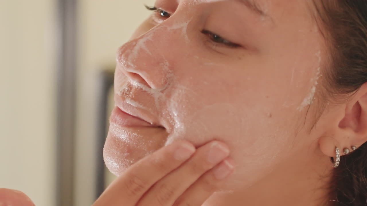 Hotel bathroom closeup woman applying cleanser gentle foam spread across face, fingers massaging forehead and cheeks, mirror reflection, earring visible, soft warm lighting, intimate skincare ritual