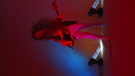 vertical video of a female bass player in a jacket jumping on one leg dancing and shaking her head and hair. crazy guitar player in the studio neon lights and spotlights