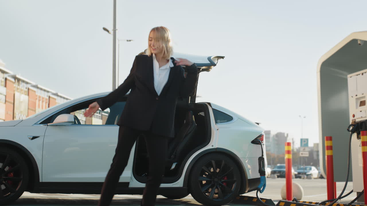 mujer feliz bailando en un coche eléctrico en una estación de carga