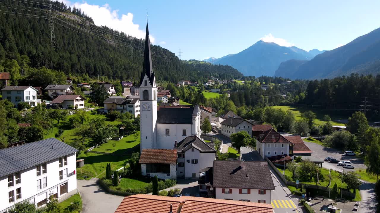 Un drone scende sopra Surava mostrando i tetti delle chiese del villaggio e la valle alpina in un'ampia vista panoramica.