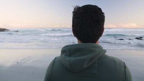 static shot of jogger in right of frame from behind pensively looking out at ocean from beach in jogging hoodie in the early morning