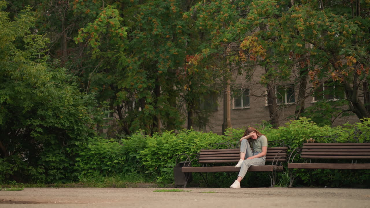 젊은 여성은 공원의 나무 벤치에 앉아, 평화로운 야외 환경에서 고독과 상을 표현하는 울창한 초록색과 나무로 둘러싸인 반사적이고 우울한 자세로 팔에 머리를 고 있습니다.