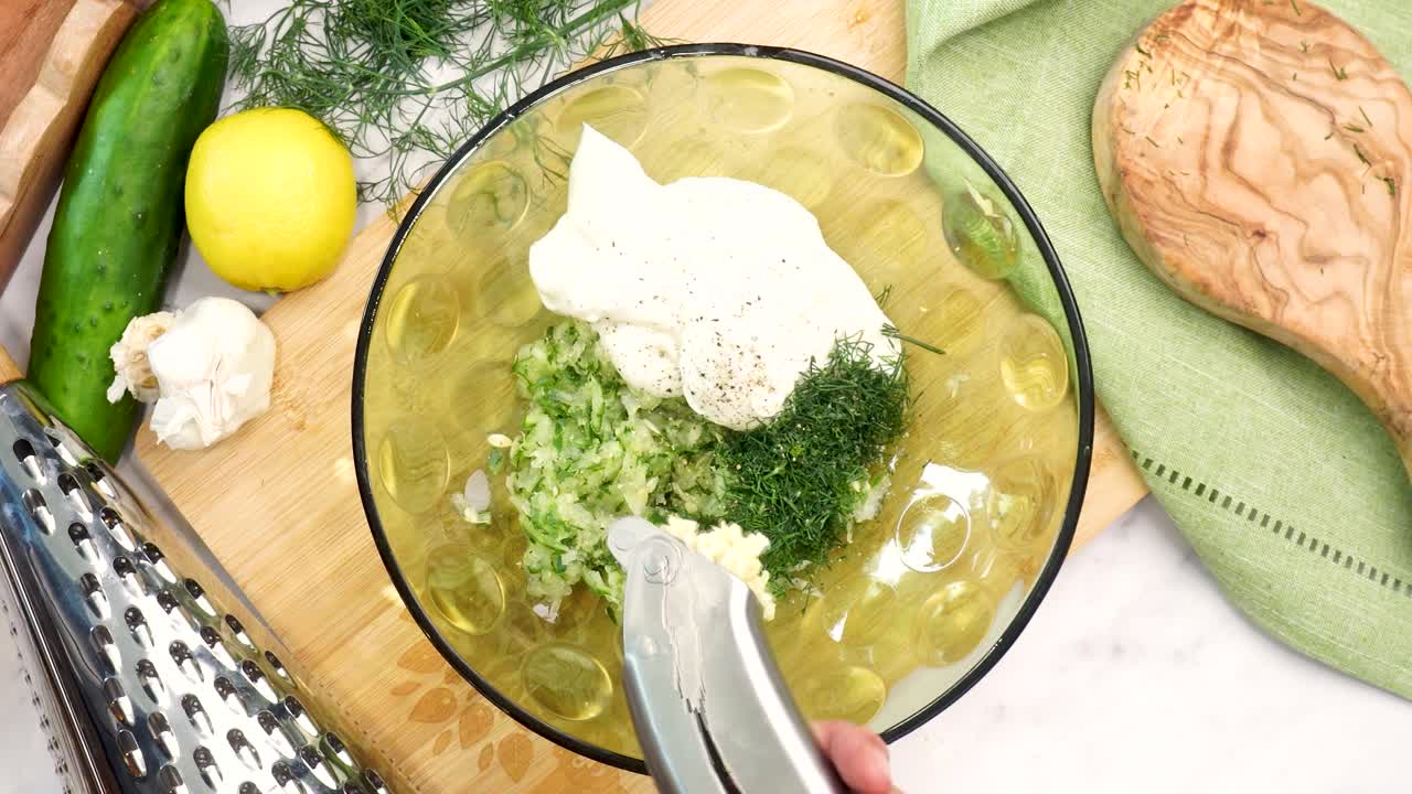 Making Tzatziki Greek or Mediterranean sauce or dip for a party. Dip or sauce in a green bowl on a wooden cutting board with dill, green napkin and a grater. Mincing garlic cloves into Tzatziki sauce.