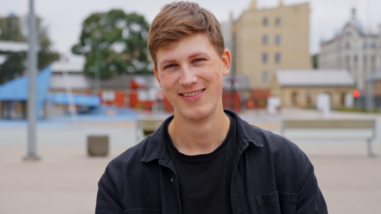 Dolly forward to smiling young white man sitting relaxed on bench in urban park, slow motion