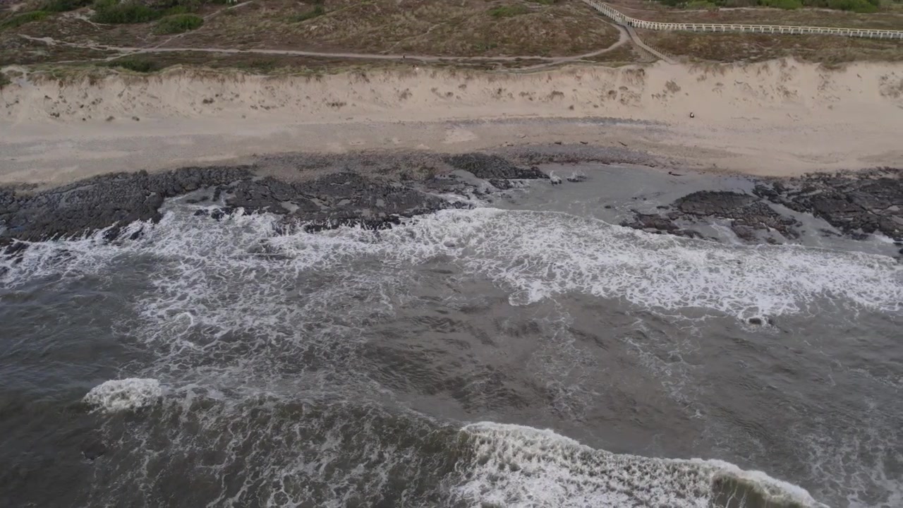 Onde che si infrangono su rocce nere su una spiaggia sabbiosa a Minho, nel nord del Portogallo, con un paesaggio di dune aspre sullo sfondo