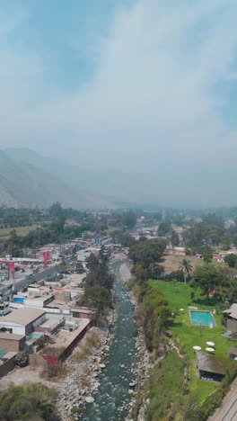 bright aerial footage of a rural city in lima, peru, captured at midday. the shot highlights a flowing river, green fields, and distant mountain peaks under the clear, sunny sky.